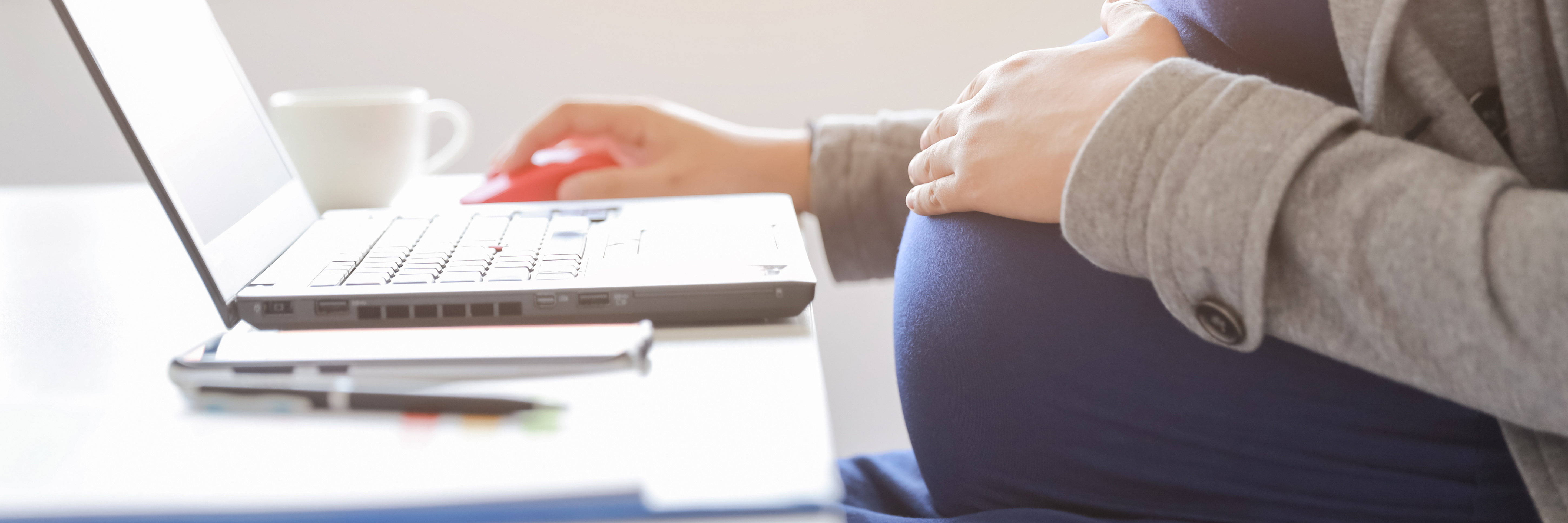 Auf dem Bild sieht man den Bauch einer schwangeren Frau, die am Schreibtisch sitzt und am Laptop arbeitet.