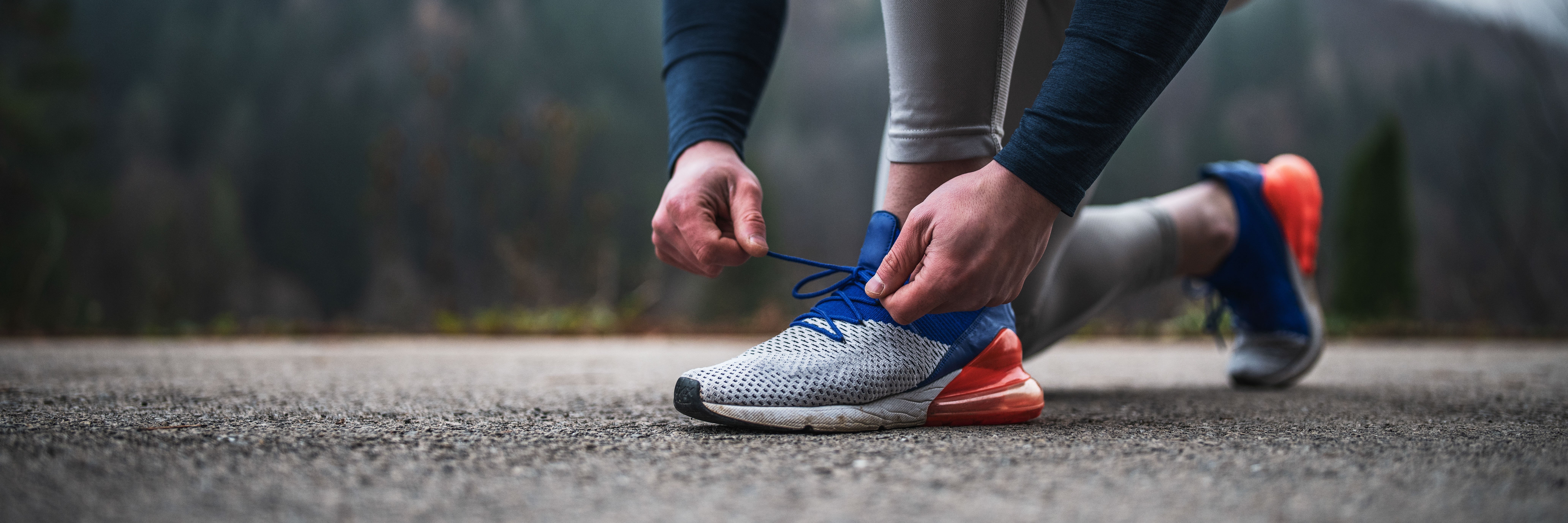 Auf dem Bild sieht man Beine und Arme einer Person in Sportkleidung, die sich gerade die Schuhe zubindet, um Laufen zu gehen. Die Person befindet sich auf einer Straße, vermutlich vor einem Waldstück.
