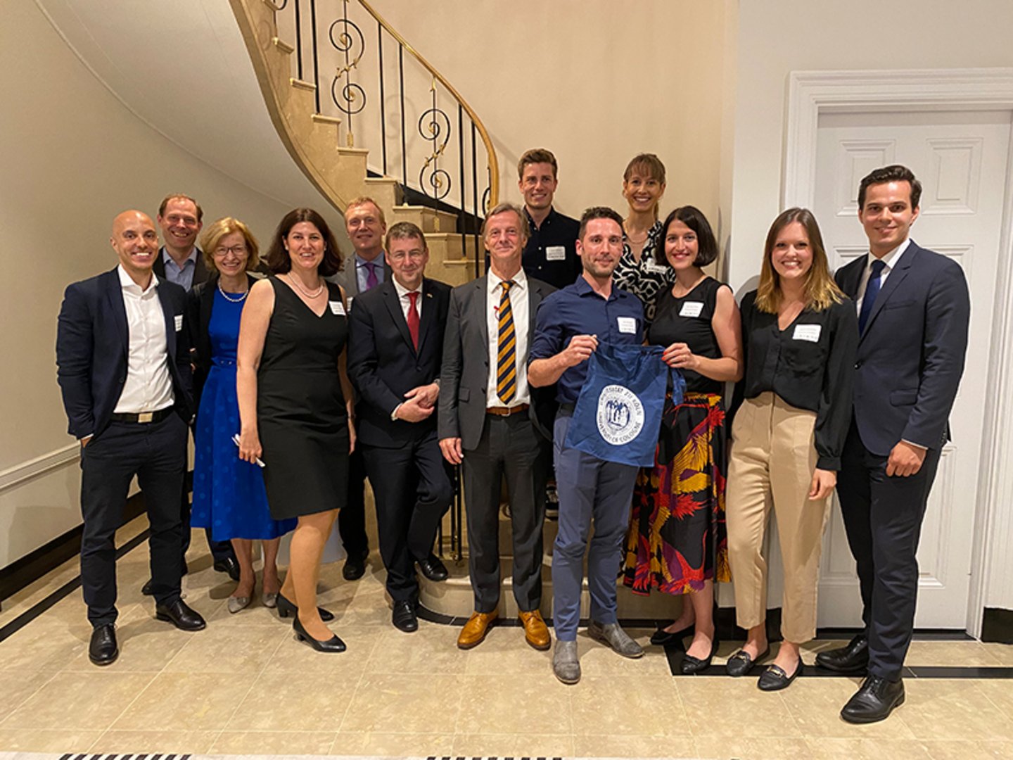 Group of people standing by a staircase, one holding a University of Cologne tote bag
