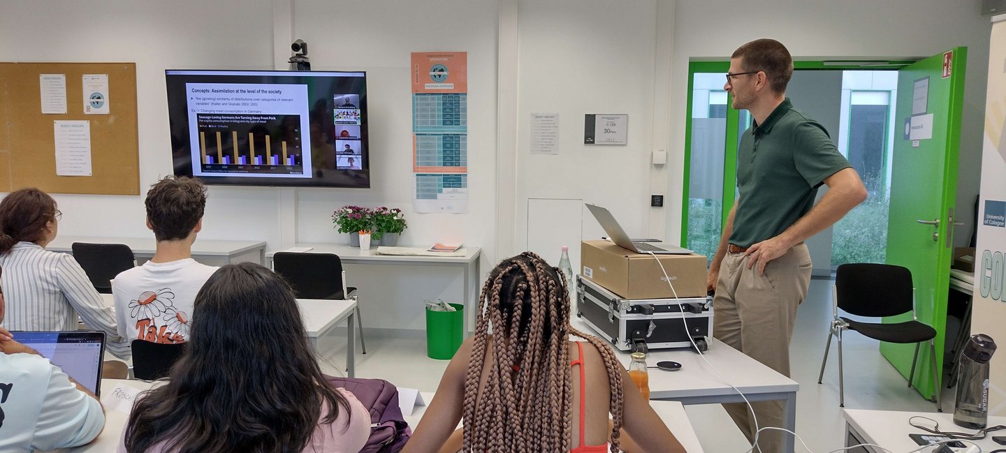 the hybrid seminar room of Cologne Summer School. During the lecture of Prof. Kroneberg (UoC) onsite and online students look at his slides and ask questions.