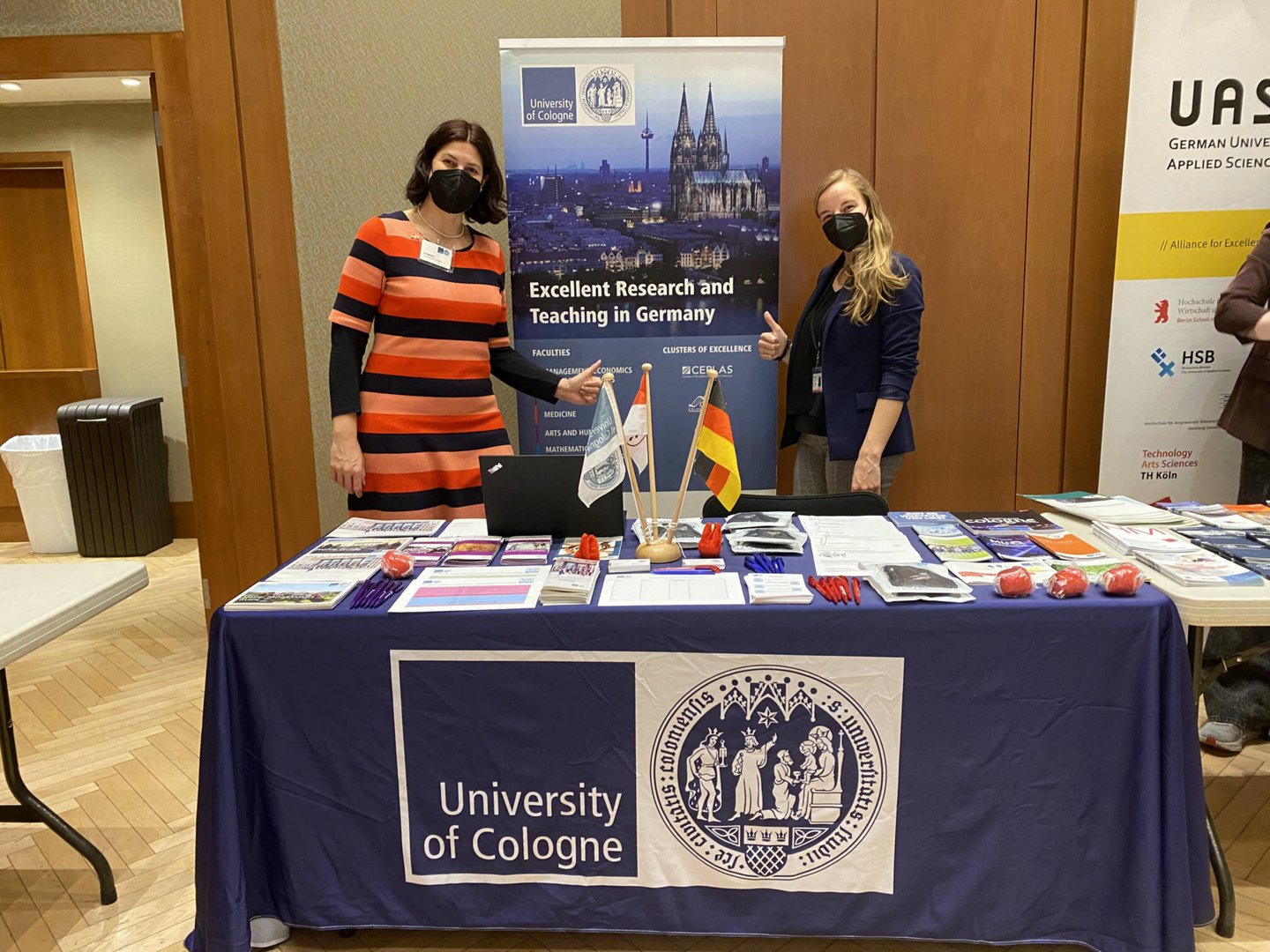 Two masked women standing at a University of Cologne booth