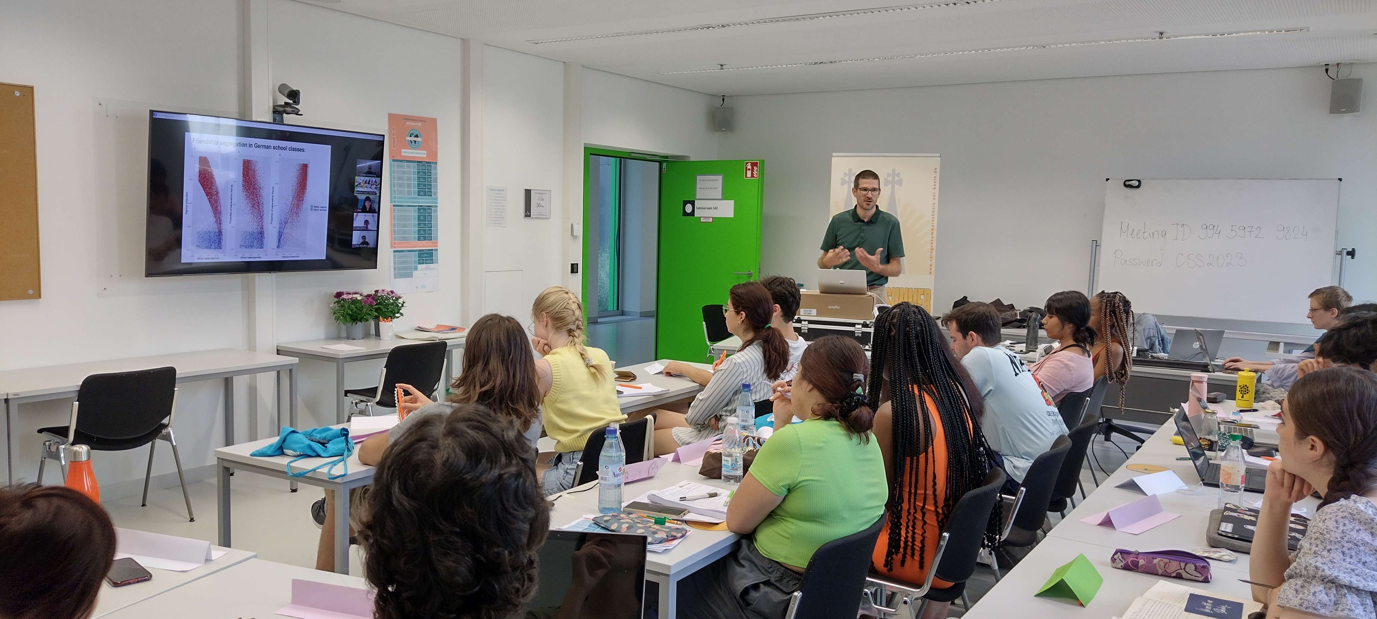 the hybrid seminar room of Cologne Summer School. During the lecture of Prof. Kroneberg (UoC) onsite and online students look at his slides and ask questions.